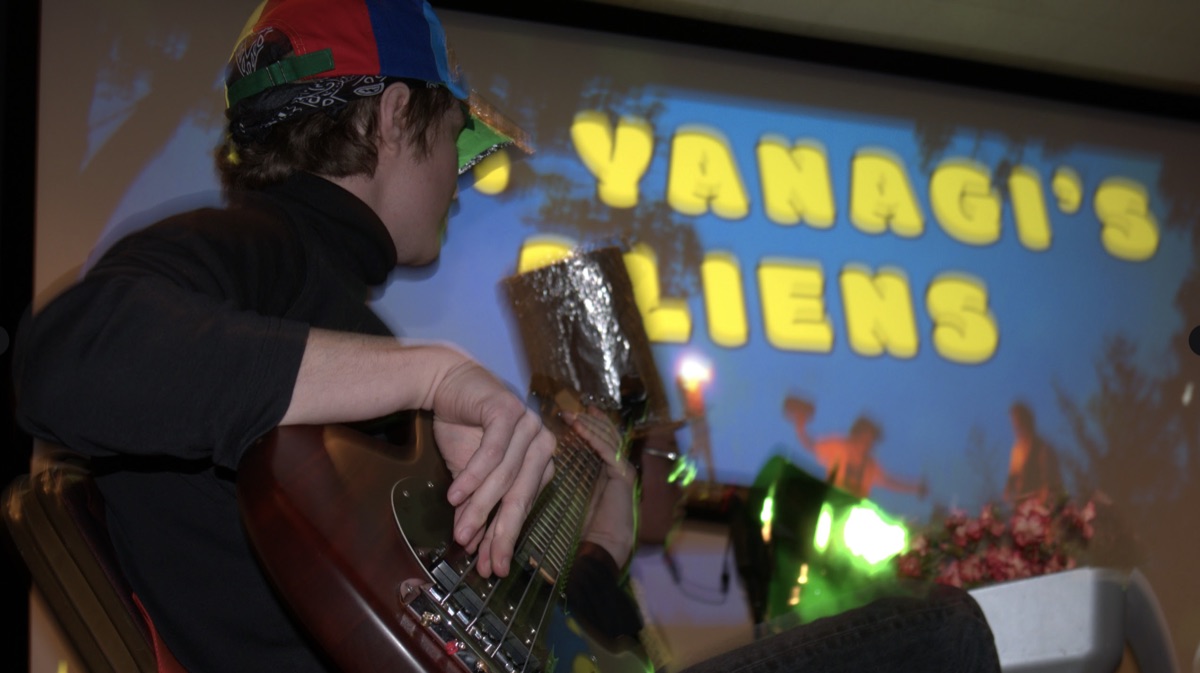 Silhouette of a guitarist performing in front of a projection reading 'Yanagi's Aliens', from the Mr. Yanagi's Aliens premiere