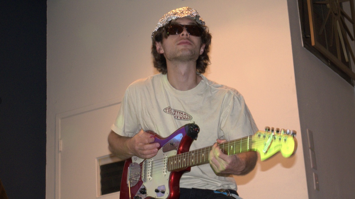A musician playing a red and green electric guitar while wearing a tinfoil hat, from the Mr. Yanagi's Aliens premiere