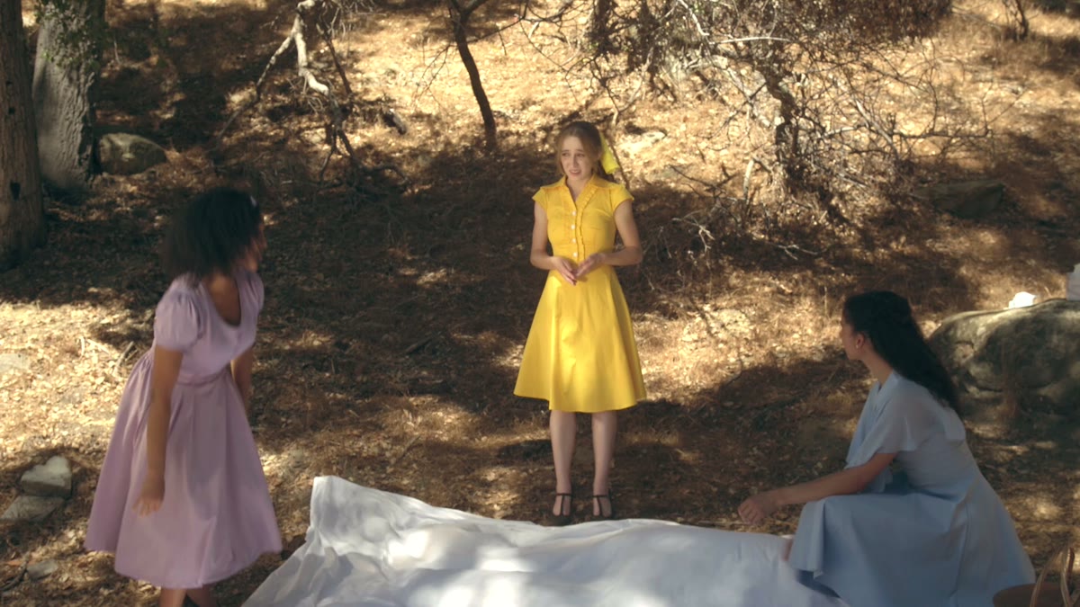 Three women in vintage dresses standing in a sun-dappled forest, a still from Left Hook Ladies Club